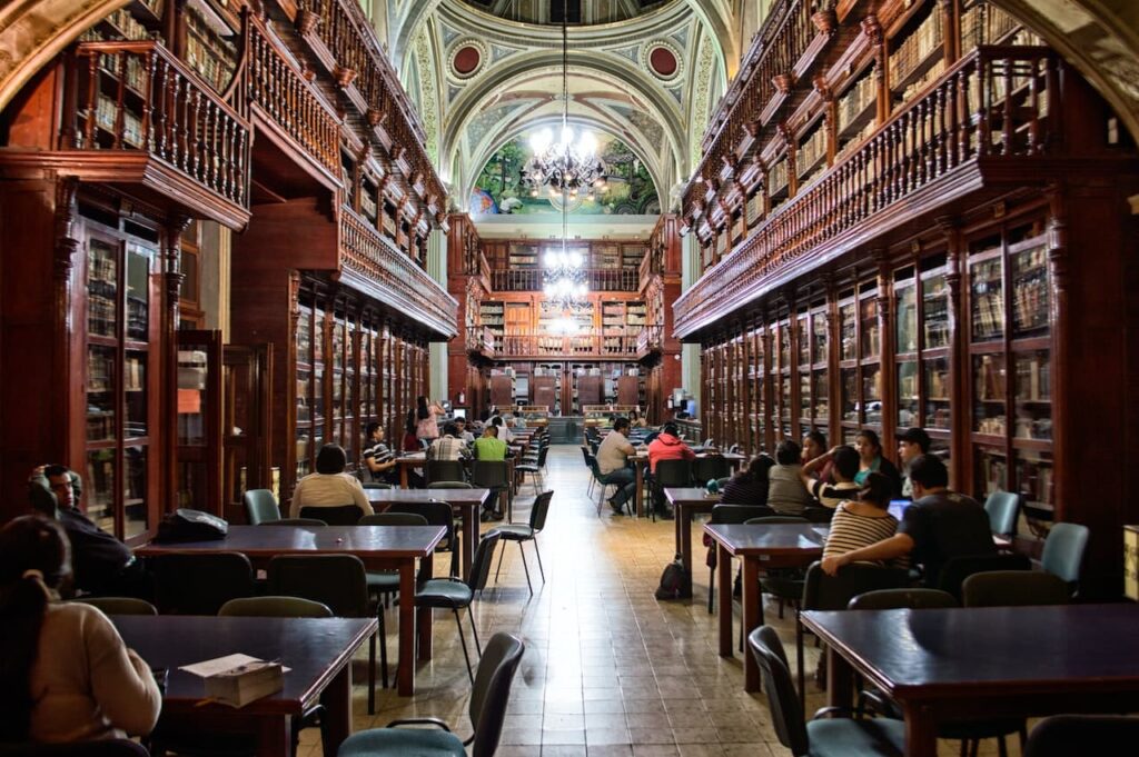 Biblioteca antica con pareti in legno e corridoio illuminato da lampadari appesi a un soffitto rinascimentale a volte. A sinistra e a destra ci sono tavoli e studenti che studiano.