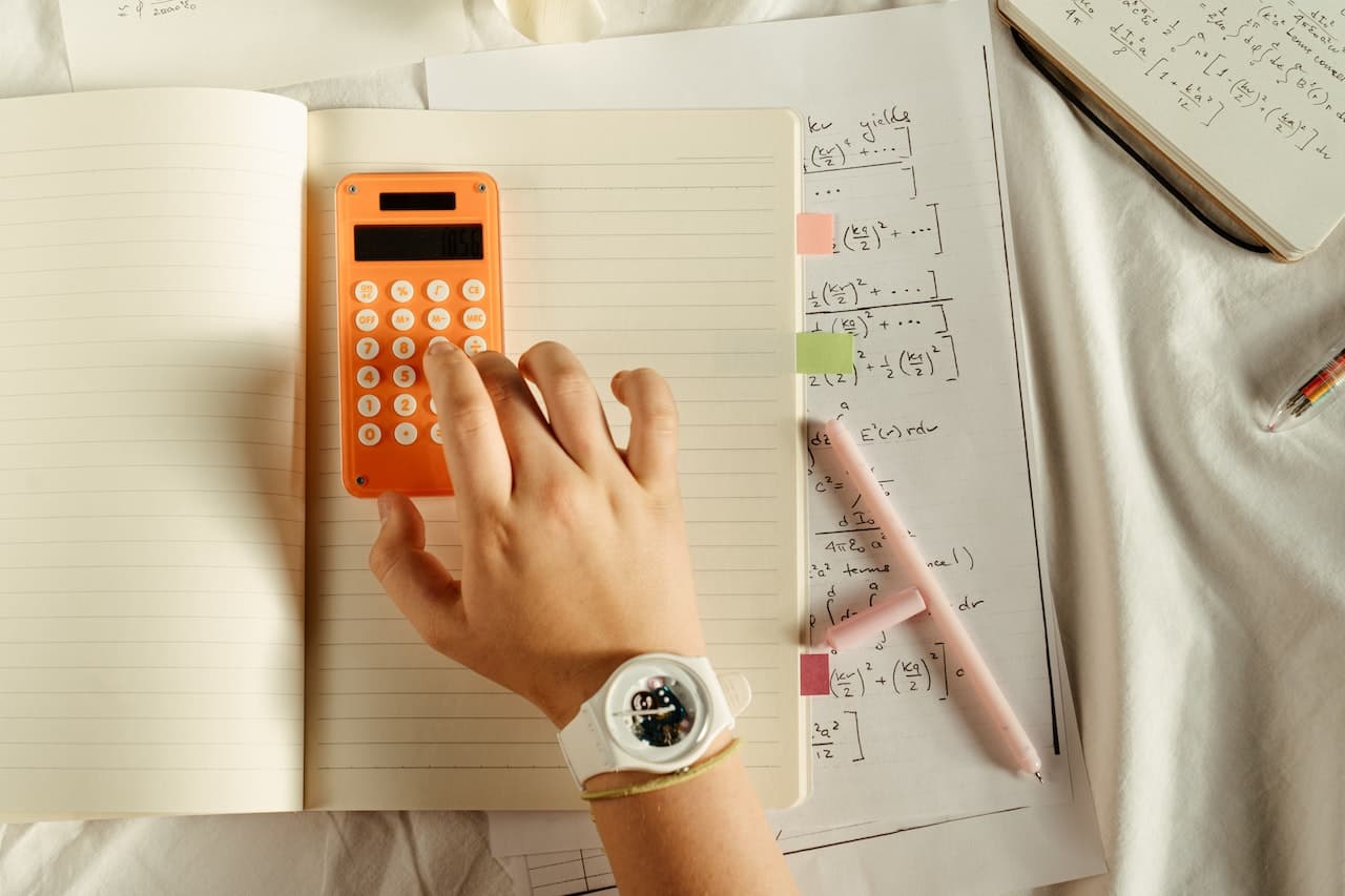 Hand with white watch on wrist while typing numbers on an orange calculator to carry out exercises on harmonic motion and its formulas.  In the background there are notebooks and pens with mathematical equations and numbers.