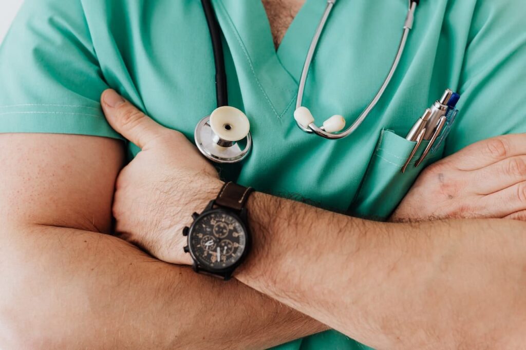 Medico con le braccia conserte. Indossa uno scrub verde, un orologio nero e uno stetoscopio al collo.