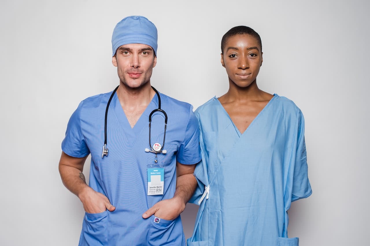 Foto frontale di due operatori sanitari in posa, indossano la divisa ospedaliera e gli accessori tipici della professione.