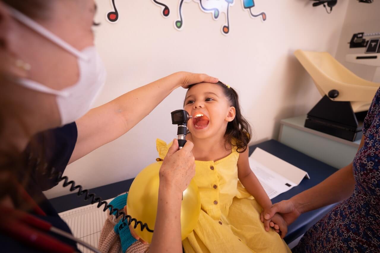 Pediatra che visita una bambina con uno strumento specifico per i controlli pediatrici. La mano del dottore è sulla testa della bambina, che è seduta sul lettino con vestito giallo e lingua fuori.