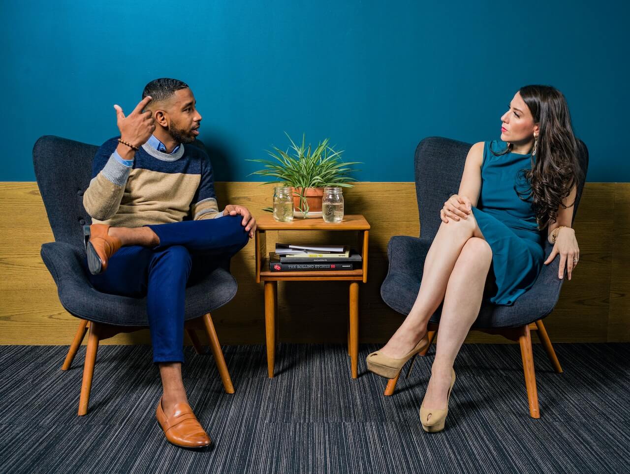 Logopedista mentre discute con un paziente per monitorare l'efficacia della terapia. I due sono seduti su delle poltrone in tessuto in una stanza con pareti blu e legno.
