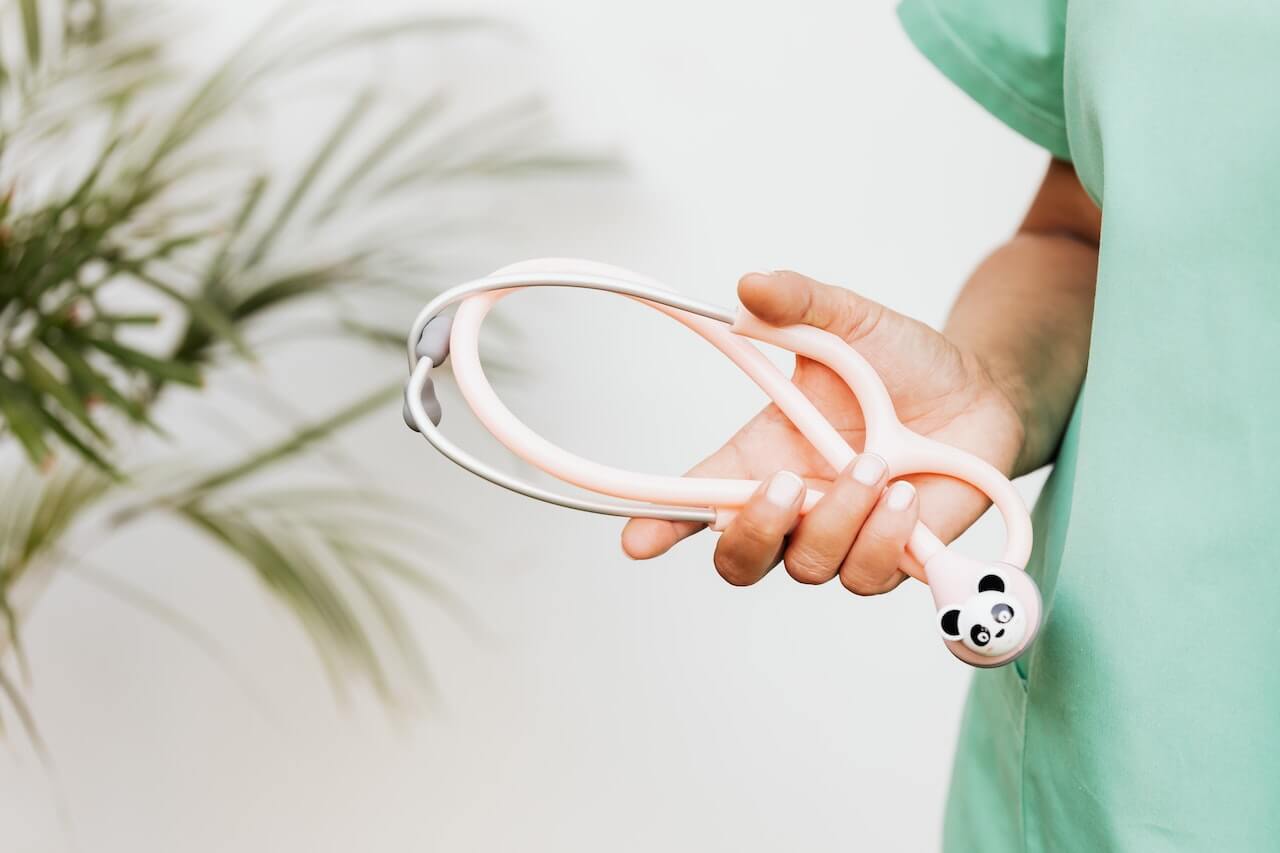 Logopedista con stetoscopio rosa e dettaglio di un panda. Il soggetto indossa uno scrub verde e sullo sfondo c'è una pianta verde sfocata.