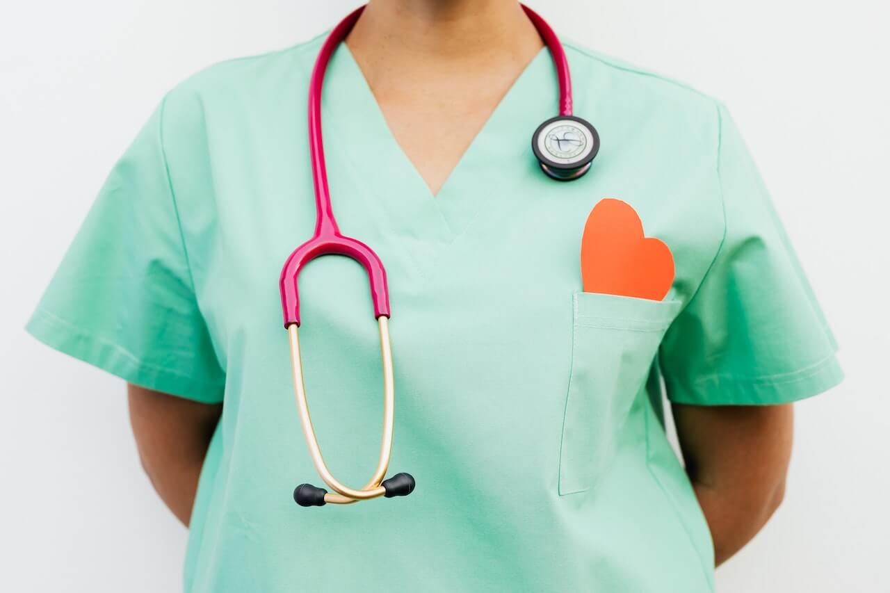 Lavoratrice delle professioni sanitarie con stetoscopio rosso al collo. Indossa uno scrub verde e ha un cuore ritagliato nel taschino.