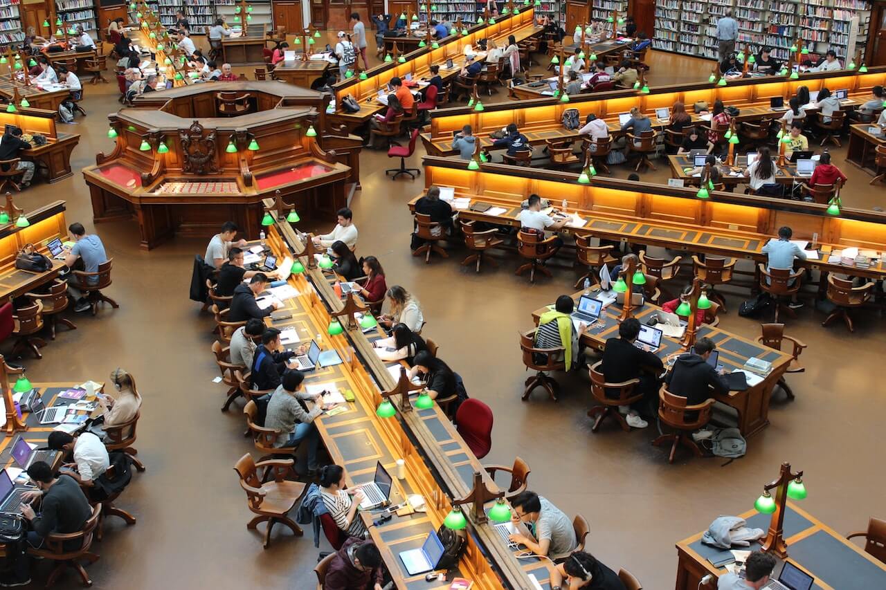 Biblioteca con tavoli lunghi e tanti studenti seduti mentre studiano e leggono.