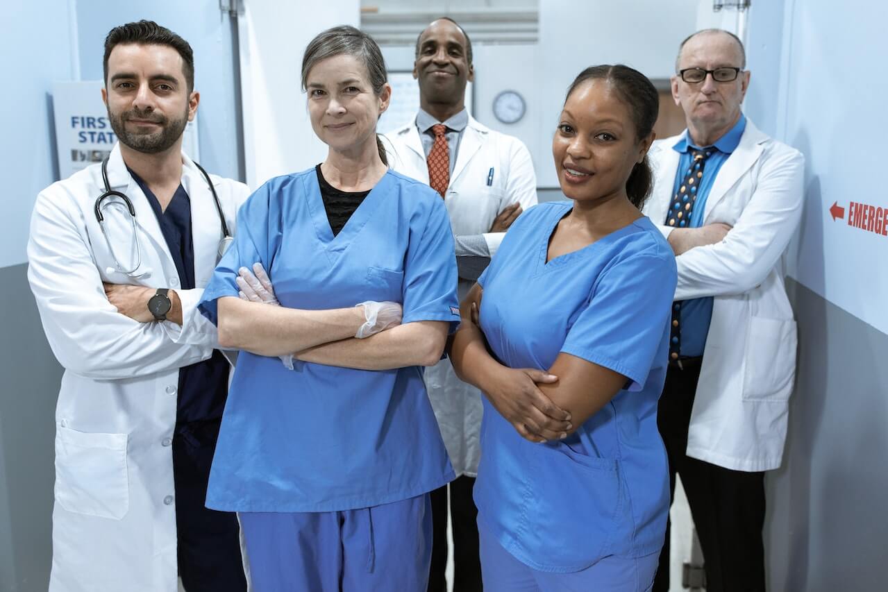 Medici e infermieri fotografati nel corridoio di un ospedale. In foto ci sono cinque soggetti, due indossano lo scrub azzurro e tre indossano il camice bianco. Il corridoio è celeste e sulla destra c'è una scritta rossa.