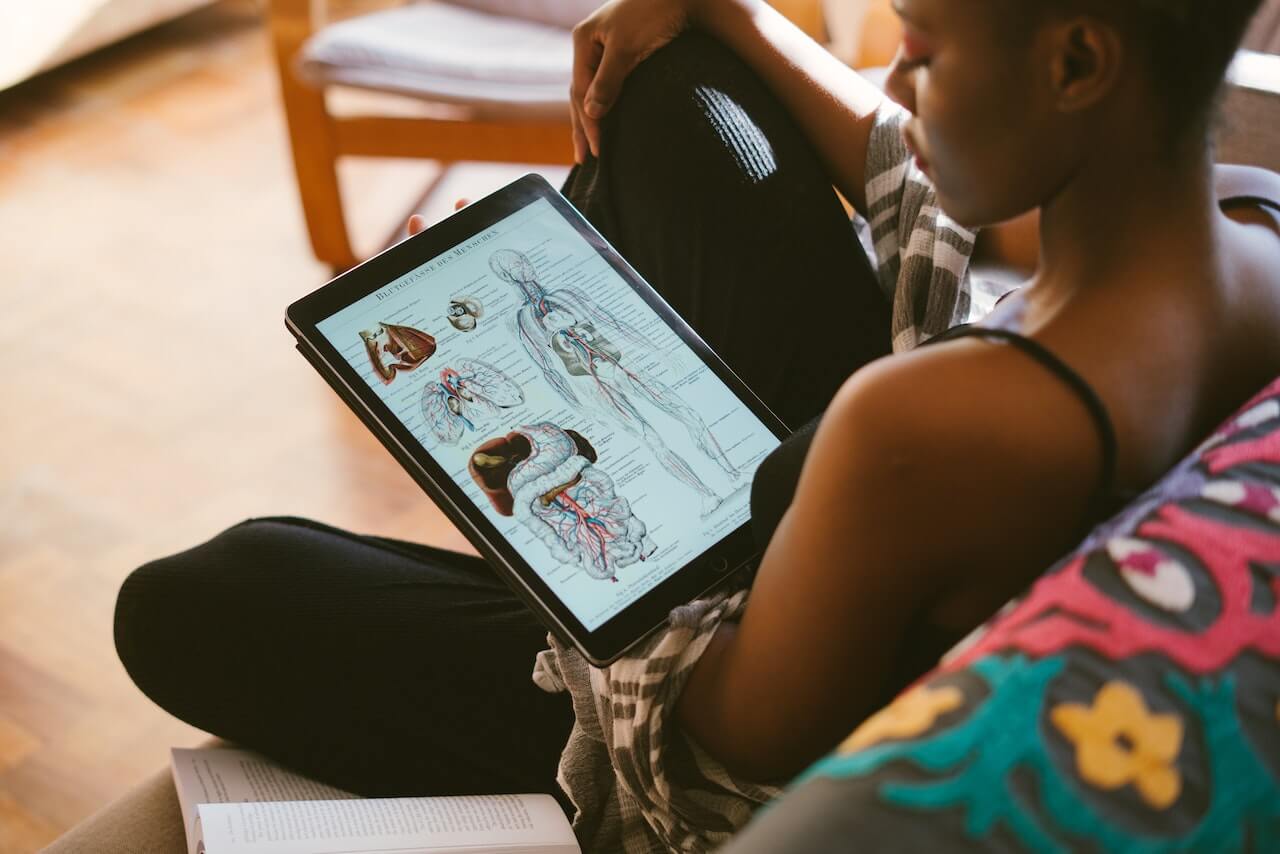Studentessa mentre legge degli appunti di anatomia su un tablet. È seduta sul divano e vicino alla gamba sinistra ha un libro aperto.