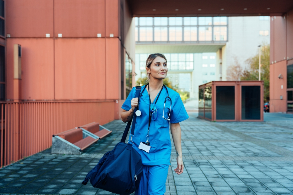Studentessa di Endocrinologia con indosso uno scrub blu e uno stetoscopio intorno al collo mentre cammina in una zona universitaria e ospedaliera.