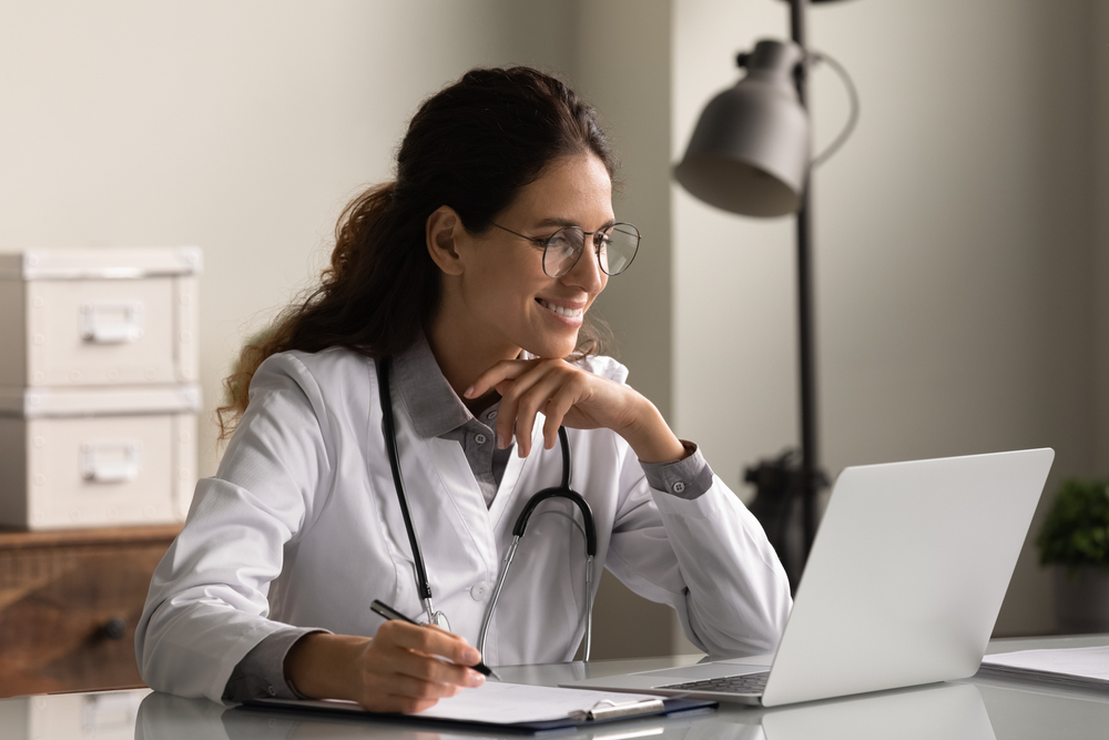 Un medico donna con occhiali e uniforme che riempe documenti sulla storia clinica del paziente, guarda lo schermo del laptop.